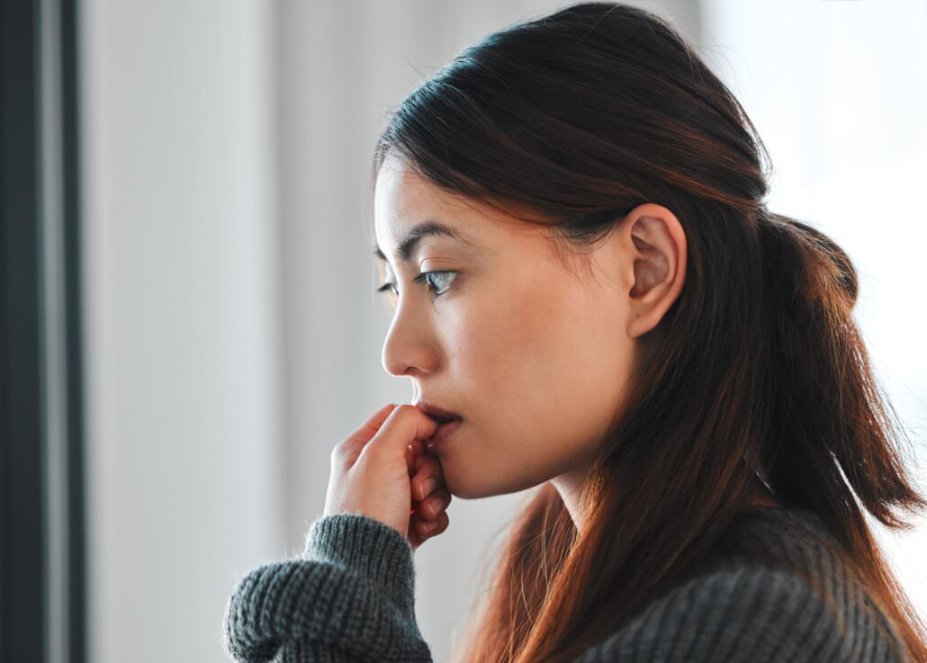 Female veteran's side profile near a window with hand to mouth and focused anxious gaze.