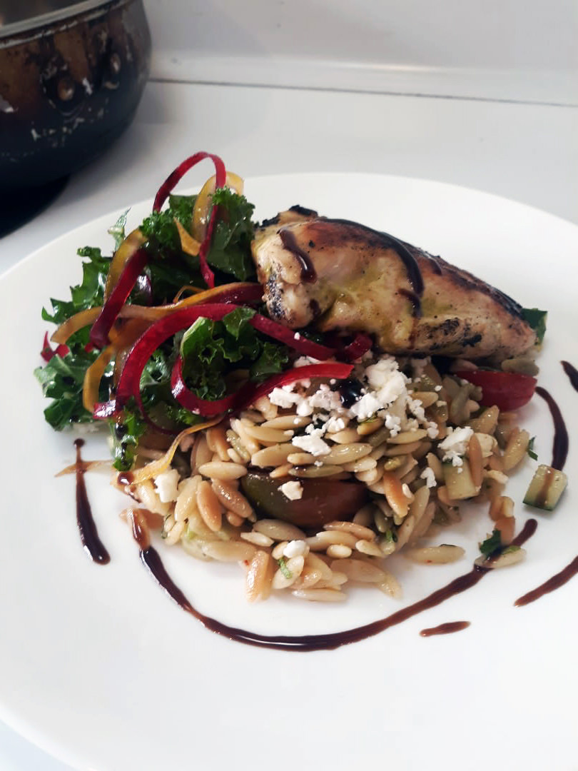 Grilled chicken breast with balsamic drizzle over kale-ribbon salad and herbed orzo on a white plate.