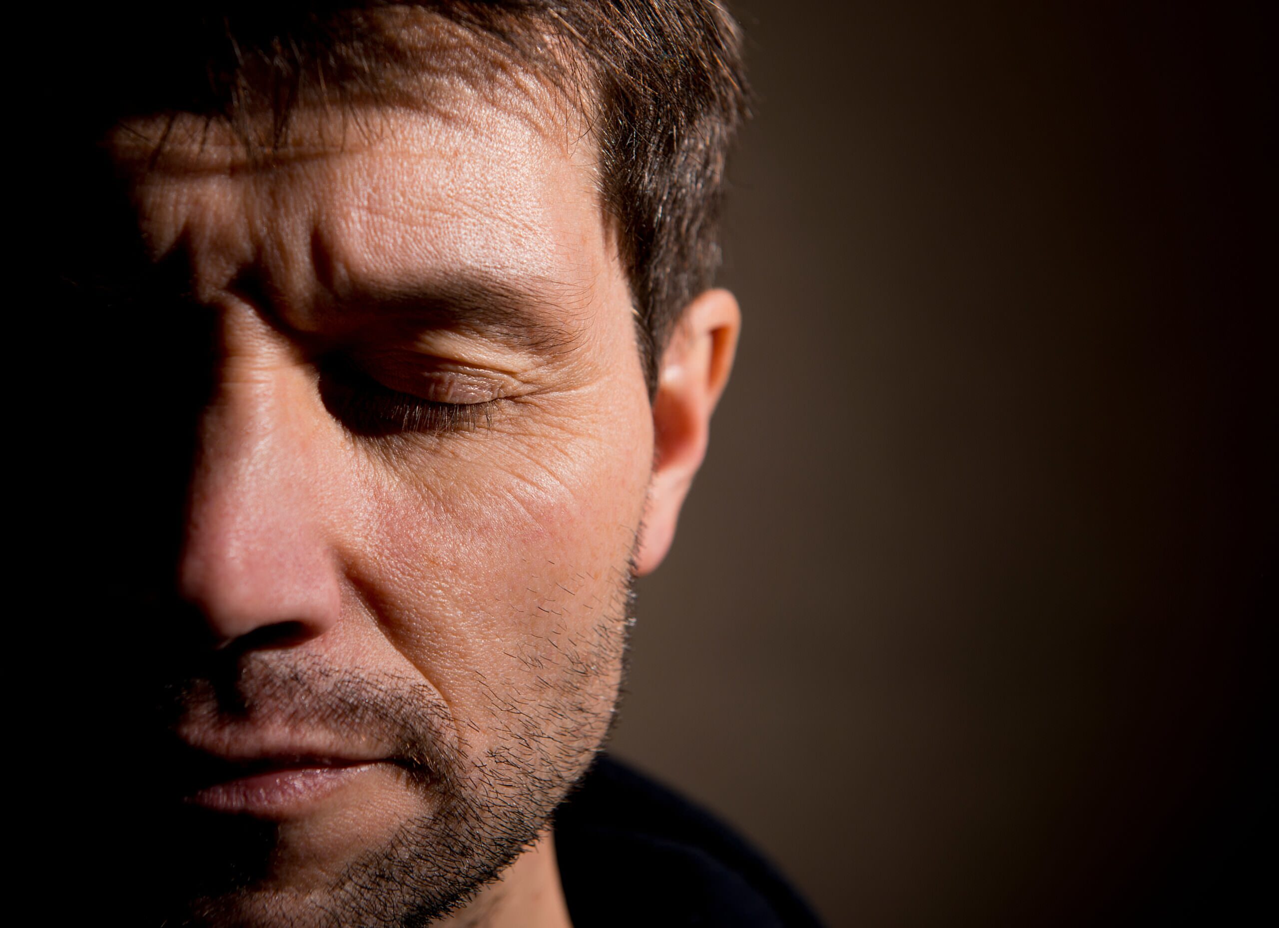 A close-up of a middle-aged man's face, half in shadow, with his eyes closed and a pained, somber expression.