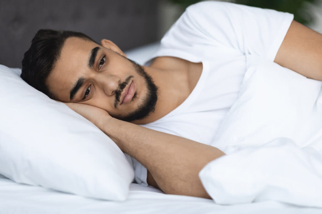 A young man with a beard lies awake in bed with his eyes open, looking troubled and unable to sleep.