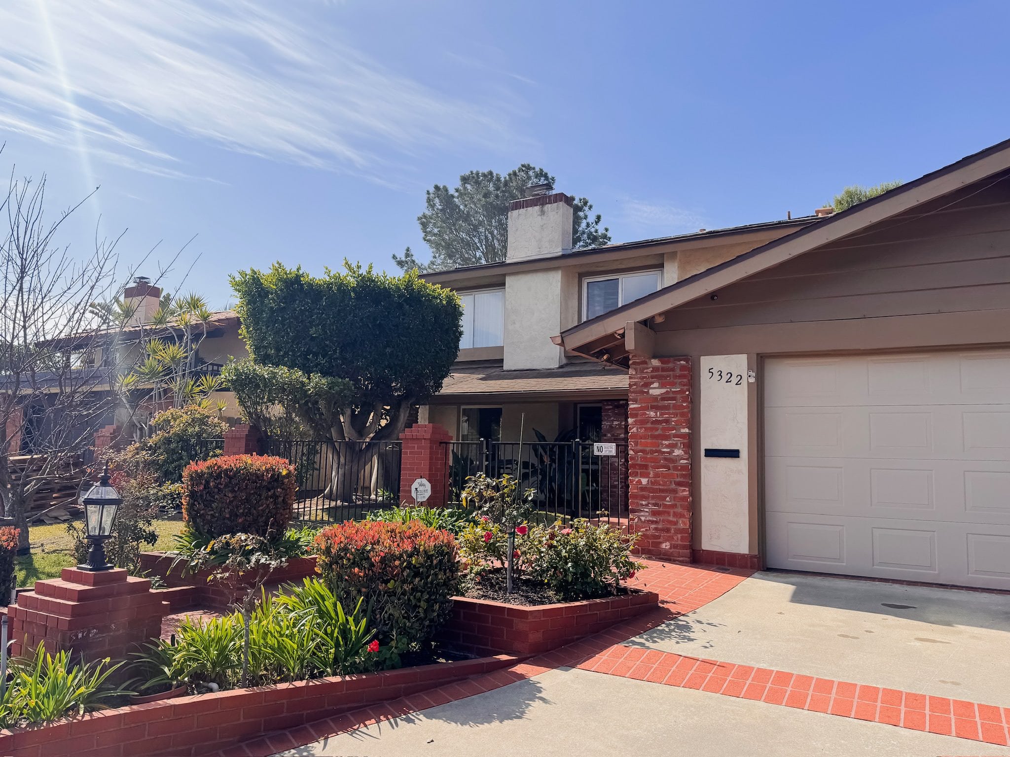 A suburban two-story house with a mix of brick and stucco exterior, red tile walkway, manicured hedges, and a front courtyard garden. The house number 5322 is displayed next to the garage.