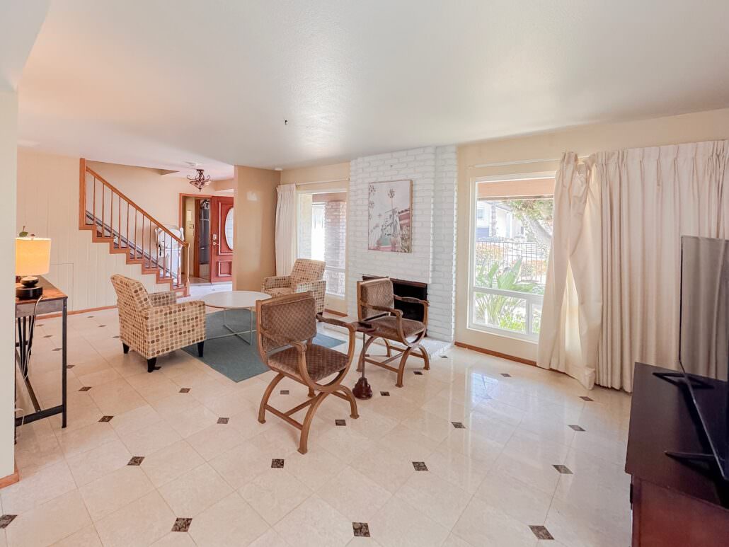 A sunlit living room with light tile floors, a white brick fireplace, and neutral-toned furniture. Large windows overlook the garden, and a staircase with a wooden handrail leads upstairs.