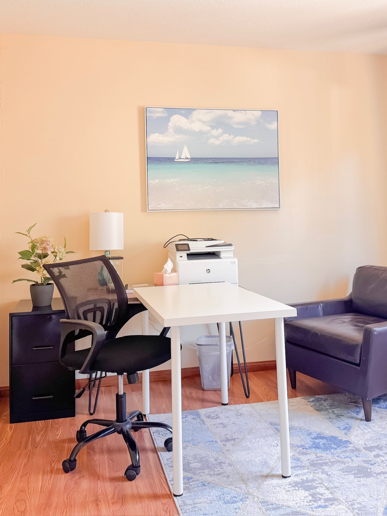 A small, tidy office with a white desk, black mesh office chair, printer, and side cabinet. A beachscape painting hangs on the wall above a light blue area rug.
