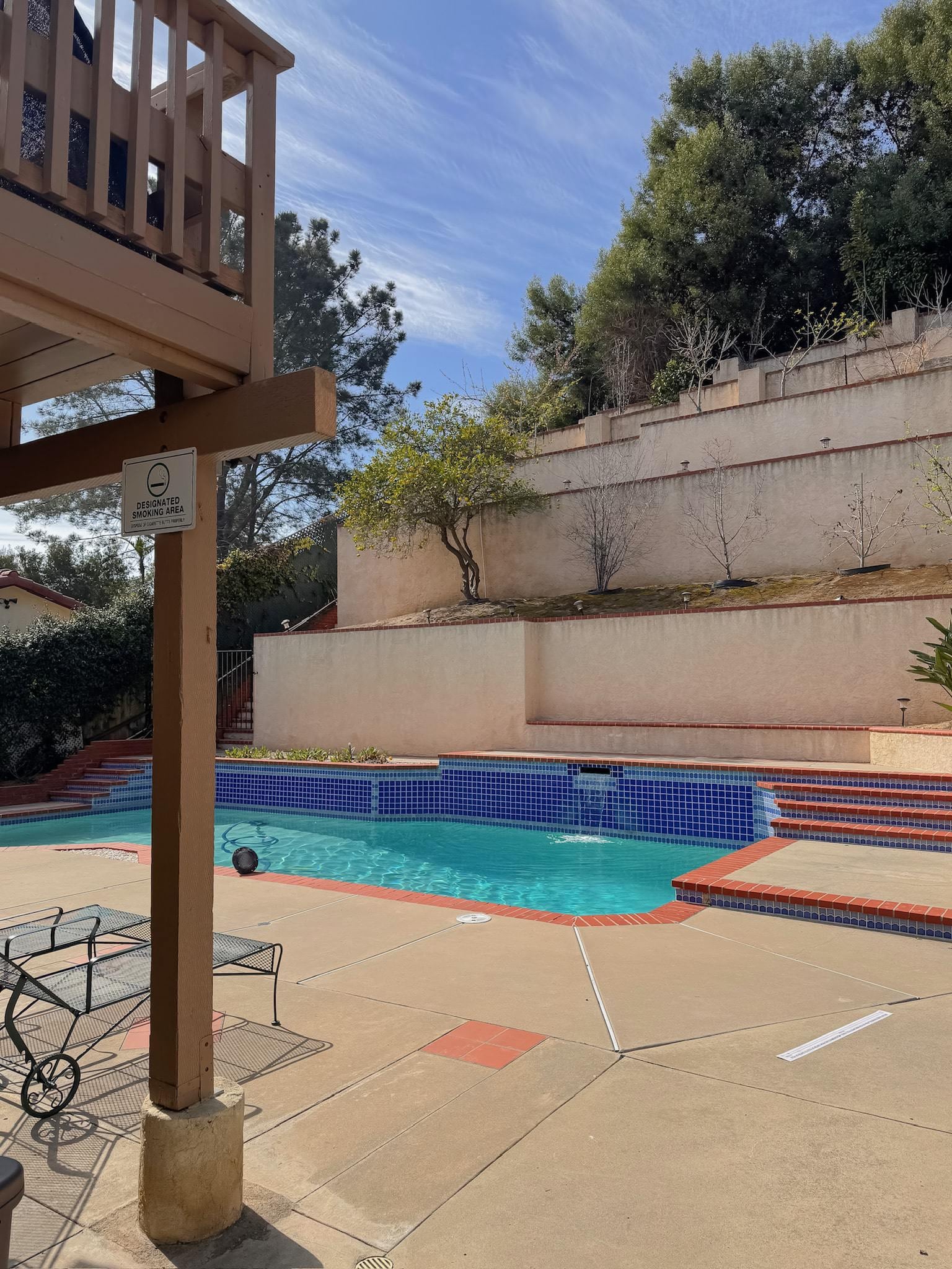A backyard pool area with clear water, red tile accents, and terraced garden walls. Surrounded by concrete decking and partial shade from a wooden patio structure.