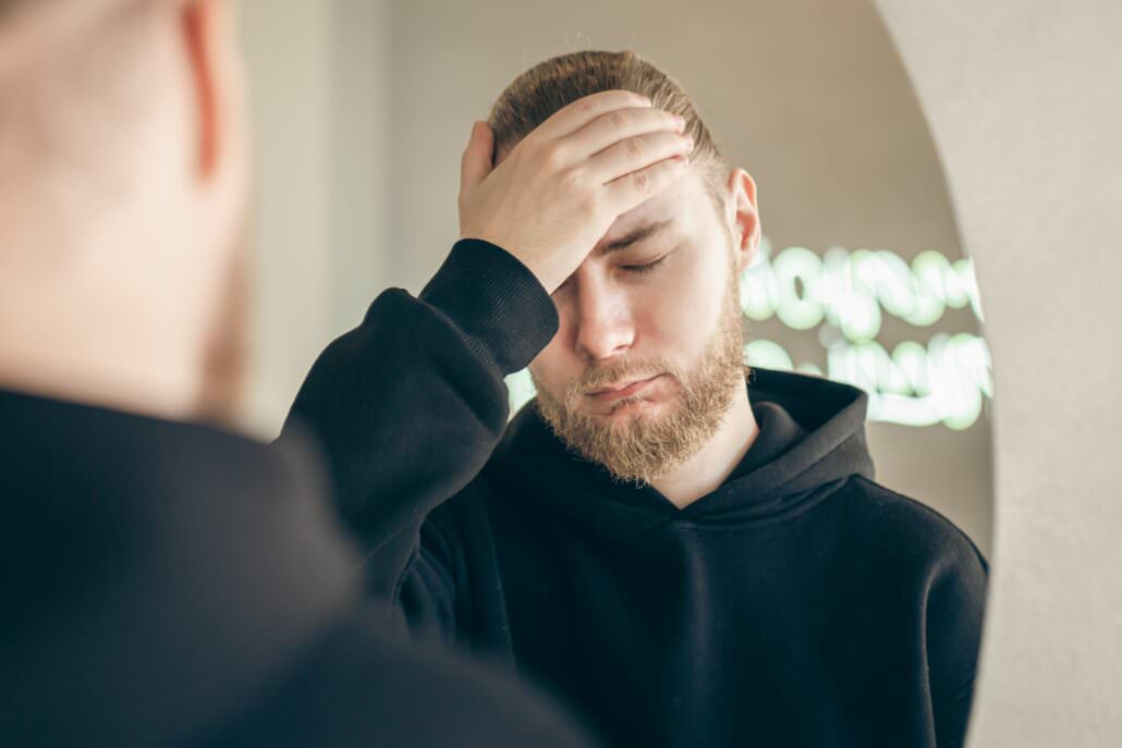 Bearded veteran in a black hoodie touching forehead beside a mirror with eyes closed.