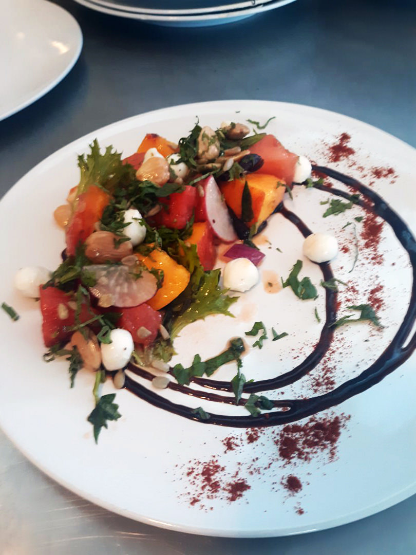 Watermelon–peach salad with mozzarella pearls, toasted seeds, herbs, and balsamic reduction swirl.