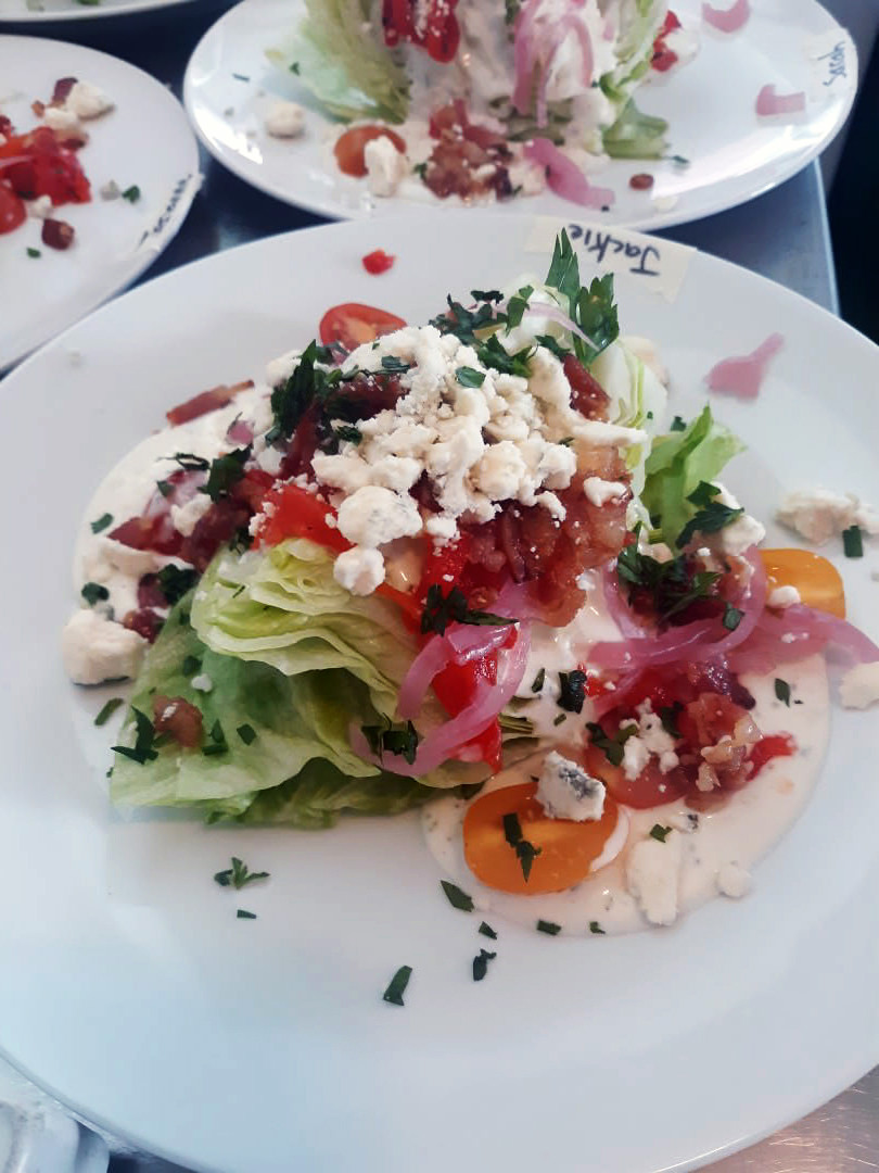 Iceberg wedge salad topped with cherry tomatoes, bacon, pickled onions, blue-cheese crumbles, ranch.